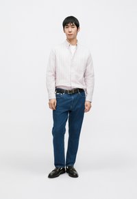 Lys rosa stripete skjorte med knapper, blå jeans, svart belte, svarte sko, står mot en ensfarget hvit bakgrunn.