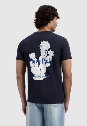 Mann von hinten, trägt ein marineblaues T-Shirt mit weißem Blumenmuster und blauem Text "Pure Path" sowie motivierenden Sprüchen auf der Rückseite.
