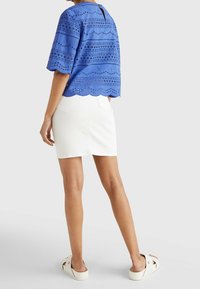 Top blu in pizzo con orlo scallopato, maniche corte e chiusura sul retro, abbinato a una gonna di denim bianca e scarpe chiare.