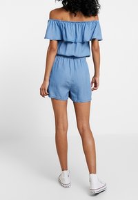 Denim off-shoulder romper i ljusblått med volangkrage, elastisk midja och korta shorts; kombinerat med vita låga sneakers.