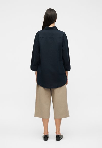 Γυναίκα με μακριά σκούρα μαλλιά που φοράει ένα φαρδύ σκούρο μπλε πουκάμισο με γυρισμένα μανίκια και μπεζ φαρδιά παντελόνια cropped, εμφανίζεται από πίσω.