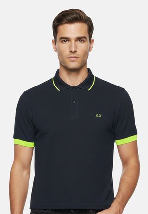 Uomo che indossa una polo nera con bordi verde neon su colletto e maniche, e un piccolo logo verde neon sul petto.