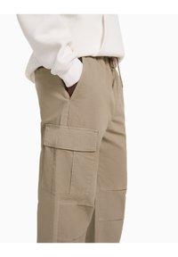 Person in beigen Cargo-Hosen mit Seitentasche und weißem Langarmoberteil, mit teilweise in der Tasche befindlicher Hand, von der Taille bis zu den Knien gezeigt.