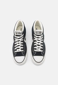 Sneakers neri in tela con suole in gomma bianca, dotati di lacci bianchi e un'etichetta sulla linguetta che dice "Converse All Star." Design classico alto.