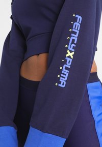 Granatowy top z długim rękawem z kontrastującymi niebieskimi rękawami, z nadrukowanym logo „FENTY PUMA” w kolorach niebieskim i żółtym na lewym ramieniu. Miękka faktura tkaniny.
