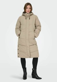 Abrigo acolchado beige con capucha, que presenta un diseño acolchado, cierres a presión y un largo considerable. Combinado con pantalones ajustados negros y botas de tobillo.