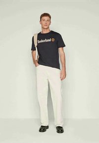 Camiseta de algodão preta com o logótipo "Timberland", combinada com calças beijas claras e sapatos de pele pretos. O modelo está de pé contra um fundo simples.