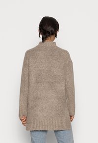 Beige Strickpullover mit hohem Kragen, strukturiertem Stoff, legerem Schnitt und geripptem Saum, getragen über hellblauen Jeans.