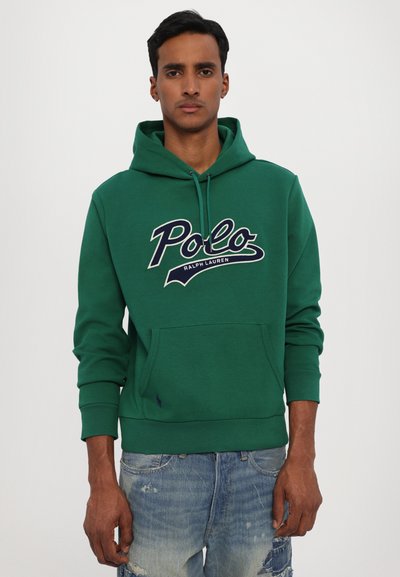 Felpa verde con logo frontale, cappuccio con cordino e tasca a marsupio. Realizzata in tessuto morbido, presenta polsini e orlo a costine. Indossata con jeans strappati.