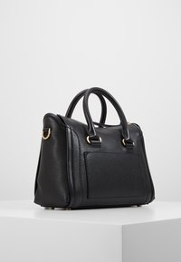 Borsa a tracolla in pelle nera con texture, dotata di doppi manici, anelli in metallo dorato e una tasca frontale, posizionata su un piedistallo bianco contro uno sfondo neutro.