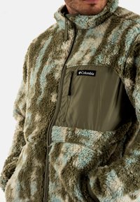 Giacca in pile verde oliva e crema con motivi azzurri chiari. Presenta una tasca sul petto con zip e un tessuto morbido e strutturato.