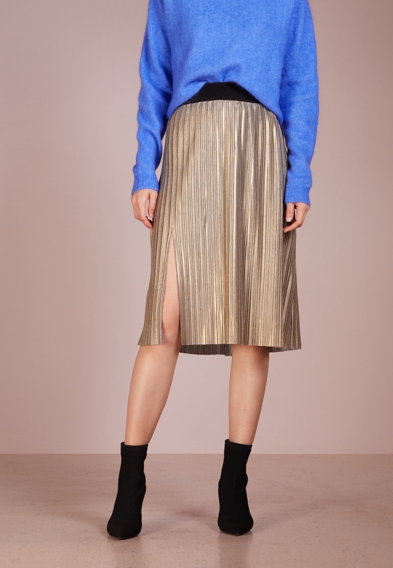 Femme portant une jupe plissée métallique longueur genou avec une fente latérale, un pull bleu et des bottines noires, debout sur un sol neutre.