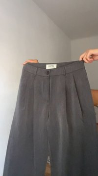 Graue Leinenhosen mit einem Bund, vorne Falten und einem lila Knopfverschluss. Der Stoff hat ein weiches, strukturiertes Aussehen.