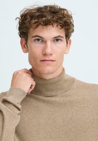 Beige garbónyi pulóver, lágy textúrával, magas gallérral és bordás mandzsettákkal. A modell egy kézzel állítja a gallért.