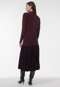 Frau, die sich abwendet, trägt ein burgunderfarbenes Langarmoberteil, einen plissierten, dunkelvelours Midi-Rock und schwarze, kniehohe Stiefel mit Absatz.