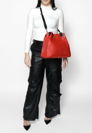 Vrouw draagt een witte coltrui, zwarte leren cargobroek, zwarte puntige schoenen en draagt een rood gestructureerde schoudertas met zwarte band.