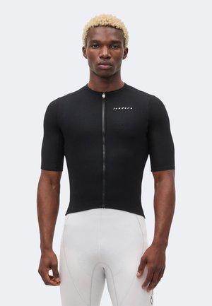 Atletisch persoon met kort blond haar draagt een nauwsluitend zwart wielertenue met rits aan de voorkant en witte gewatteerde wielerbroek.