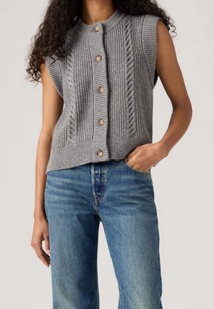 Cardigan en maille gris sans manches avec boutons devant, porté par une personne avec une main dans la poche d’un jean bleu, fond neutre.