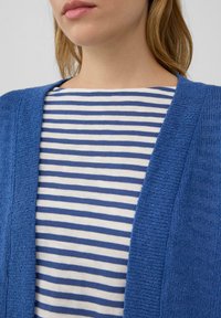 Blauwe gebreide cardigan met een gestructureerde afwerking, gedragen over een gestreepte witte en blauwe top. De cardigan heeft een V-hals en een open voorkant.