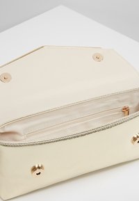 Kremowa kopertówka otwarta, pokazująca wnętrze z zamkiem błyskawicznym oraz złote zapięcia na magnes na białej powierzchni.