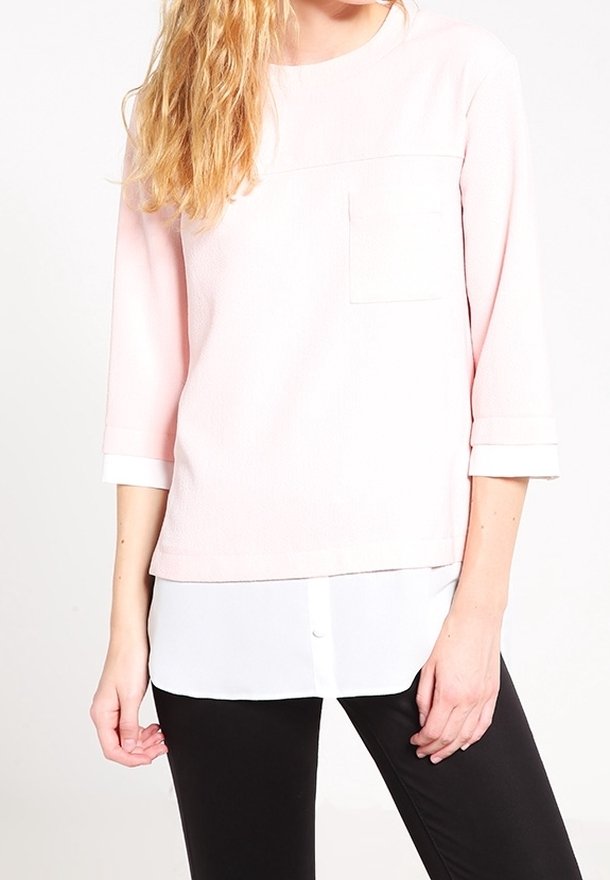 Femme portant un haut rose clair à manches trois-quarts avec une poche devant, superposé sur une chemise blanche boutonnée, associée à un pantalon noir.