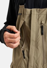 Bež vodoodporne hlače z naramnicami in zadrgo na žepu. Material se zdi gladek z mat površino, zasnovan za zunanjo uporabo.