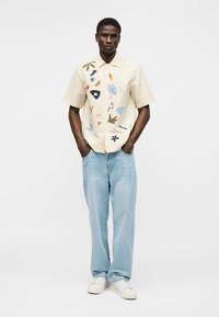 Mand iført beige kortærmet skjorte med abstrakte farverige mønstre, lyseblå jeans og hvide sneaks, står med hænderne i lommerne.