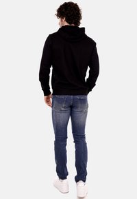 Felpa nera con cappuccio e tasca a marsupio frontale, abbinata a jeans blu aderenti. Sneakers bianche completano l'outfit.