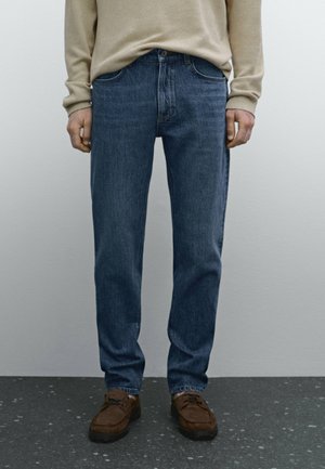 Personne portant un jean bleu, un pull beige et des chaussures en daim marron debout sur un sol sombre tacheté contre un mur clair uni.