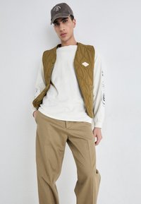 Vest olive matlasat cu închidere cu capse peste o cămașă cu manșon lung de culoare bej deschis, asortată cu pantaloni khaki; croială lejeră, casual. Șapcă gri pe cap.