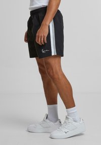 Shorts noirs avec une bande latérale blanche et bleu clair, taille élastique et un petit logo sur l'ourlet ; accompagnés de baskets blanches.