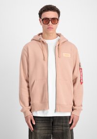 Felpa zipata rosa chiaro con tasca frontale e polsini a costine. Presenta una patch con logo sul petto e un'etichetta rossa sulla manica.