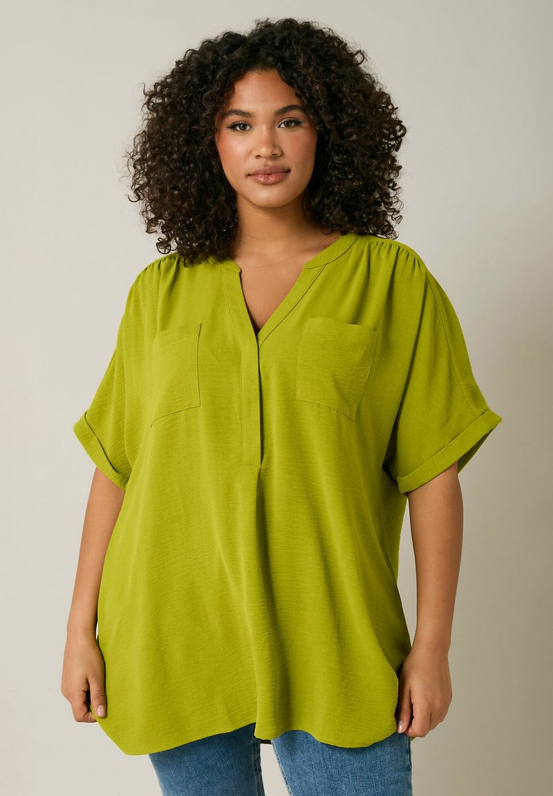 Camicia verde a maniche corte con scollo a V, due tasche sul petto, texture morbida e vestibilità rilassata, abbinata a jeans blu.