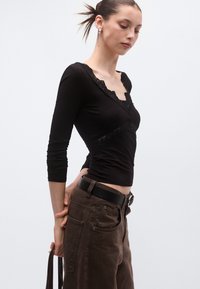 Μαύρο τοπ με μακριά μανίκια και δαντέλα κατά μήκος του ντεκολτέ, σε συνδυασμό με καφέ παντελόνι ψηλόμεσο, με υφασμάτινη υφή και ζώνη.