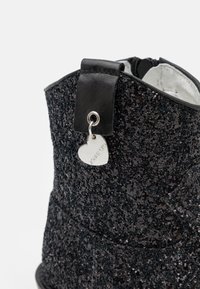 Černé třpytivé kotníkové boty s texturovaným svrškem a stříbrným přívěskem ve tvaru srdce připevněným na černé kožené smyčce. Okraje jsou hladké.