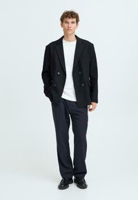Black pinstripe double-breasted blazer over a white crew neck shirt, paired with dark trousers and black lace-up shoes. Smooth fabric texture.