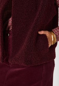 Gilet in pile bordeaux con superficie testurizzata e morbida; presenta tasche laterali ed è indossato sopra una camicetta a scacchi e una gonna in velluto a coste.