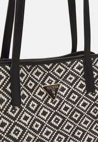 Black and white woven tote bag with leather accents, featuring a diamond pattern and a gold triangular logo plaque. Two black handles.