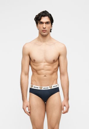 Svalnatý muž stojaci čelom dopredu, oblečený v čiernych slipoch s bielym pásom s tvárami plyšových medvedíkov a nápisom „Moschino“.