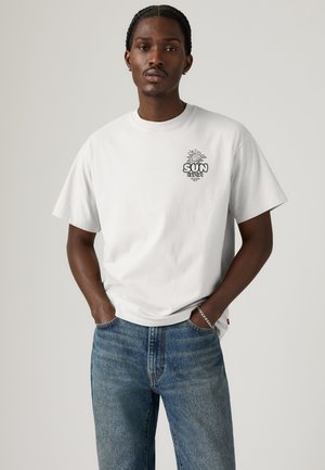 Homme aux cheveux tressés portant un T-shirt blanc avec un graphique soleil « Soul Seeker » et un jean bleu, mains dans les poches sur fond uni.