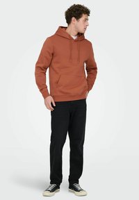 Jonge man die een roestkleurige hoodie, zwarte spijkerbroek en zwarte sneakers met witte veters draagt, staand en naar de zijkant kijkend.