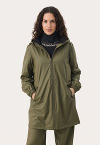 Jachetă impermeabilă de culoare verde olive cu fermoar frontal, manșete elastice și glugă. Sub aceasta se află un pulover tricotat cu guler înalt, care are un accent cu un model.