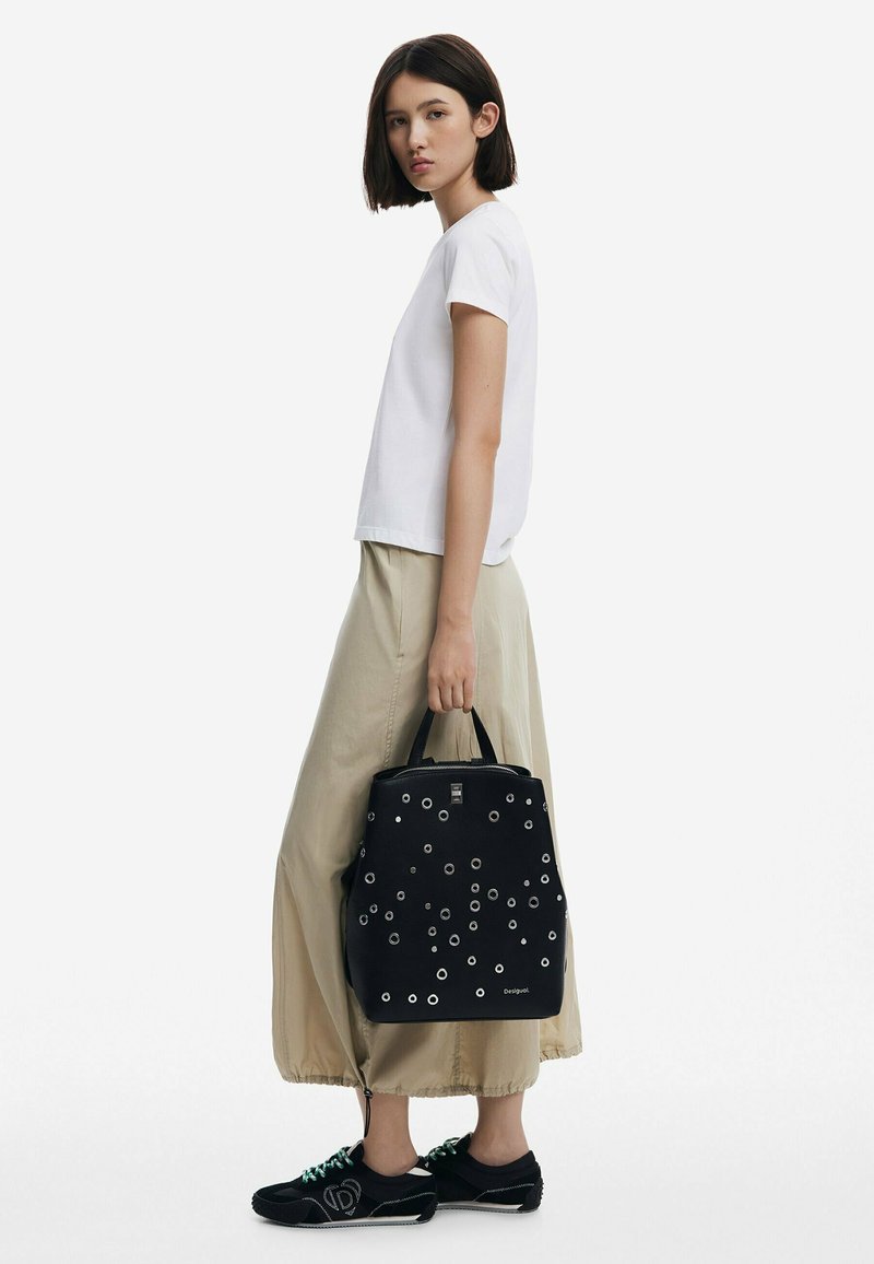 Black backpack with metallic eyelets, rectangular shape, top handles, paired with a white t-shirt and beige skirt, worn with black sneakers.