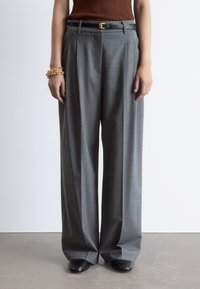 Gray wide-leg trousers with a textured fabric, pleats, and a black leather belt featuring a gold buckle. Model wears black slip-on shoes.