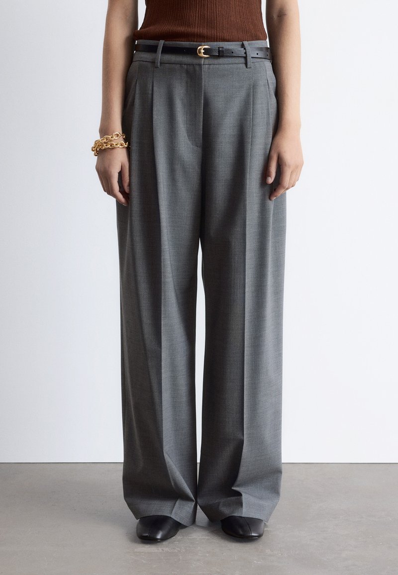 Gray wide-leg trousers with a textured fabric, pleats, and a black leather belt featuring a gold buckle. Model wears black slip-on shoes.