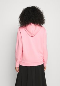 Persona con capelli ricci che indossa un felpa rosa chiaro e una gonna nera, in piedi con le spalle rivolte verso di noi su uno sfondo chiaro e uniforme.