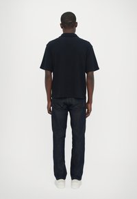 Polo shirt negru cu manecă scurtă purtat cu blugi de denim albastru închis și adidași albi; design simplu și curat, fără branding vizibil.