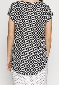 Haut à motif noir et blanc avec manches courtes, présentant un détail dos en trou de serrure. Le tissu est léger et doux, avec un ourlet arrondi.