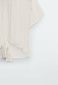 Cremefarbenes Kurzarmhemd mit Knopfleiste und einem gebundenen Knoten-Detail am Saum, aus einem weichen, leichten Stoff gefertigt.