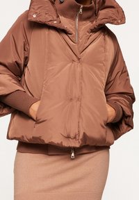 Ruskea pörrötakki, jossa on korkea kaulus, suuret hihat ja etutaskut. Sileä pinta, vetoketjusuljenta ja ribbireunat.